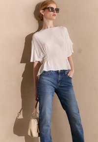 Blusa bianca ampia con arricciature laterali, abbinata a jeans blu a vita alta. Il modello tiene una piccola borsa beige con un pon-pon.