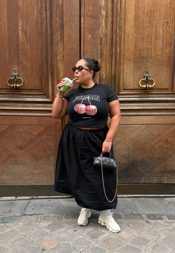 Black cropped T-shirt with a basketball graphic, black flowing skirt, white chunky sneakers, and a small black handbag. Holding a green drink.