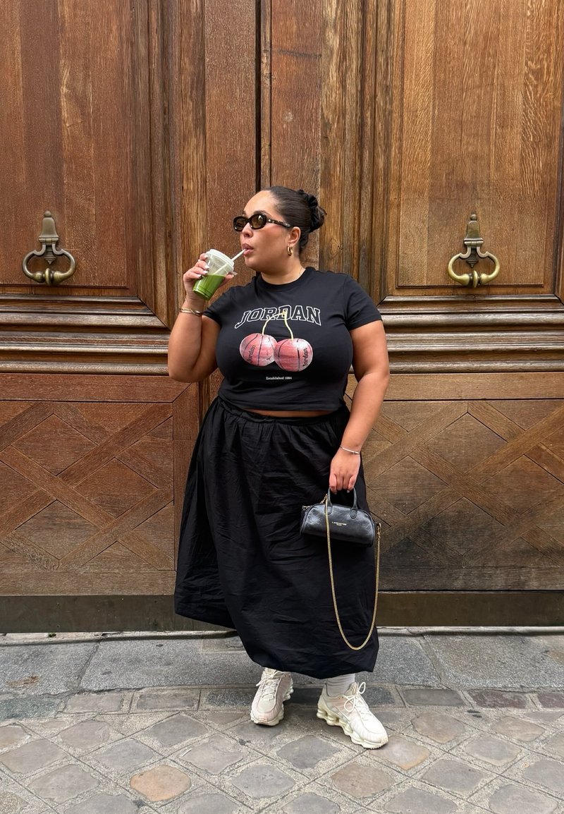 Zwarte cropped T-shirt met een basketbalafbeelding, zwarte zwierige rok, witte chunky sneakers en een kleine zwarte handtas. Groene drank in de hand.