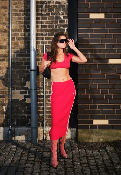 Mujer con crop top rojo y falda, botas rosas y gafas de sol negras, de pie junto a una pared de ladrillos con suelo de adoquines.