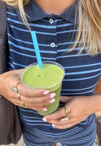 Zielony smoothie w przezroczystym kubku z jasnoniebieską słomką, trzymany w dłoniach ozdobionych złotymi pierścionkami i lakierem do paznokci. W tle widać koszulę w granatowe paski.