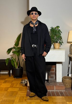 Hombre con sombrero negro, gafas de sol, pañuelo al cuello estilo bandana, atuendo negro holgado y zapatos marrones, de pie en una habitación con plantas y una chimenea.