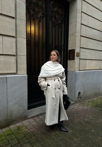 Kremo spalvos paltas su dvigubu užsegimu, diržu ant liemens ir per dideliu apykakle. Juodi aksesuarai ir iki kelių siekiančios botai puikiai papildo aprangą.