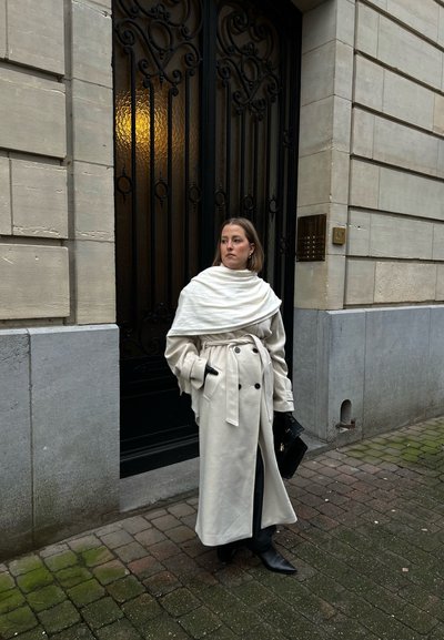 Abrigo crema con delantero de doble botonadura, cintura ceñida con cinturón y cuello sobredimensionado. Los accesorios negros y las botas hasta la rodilla complementan el conjunto.
