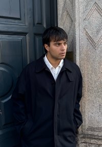 Jeune homme aux cheveux foncés, portant un manteau noir et une chemise blanche, debout les mains dans les poches devant une porte sombre et un mur de pierre.
