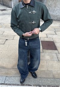 Jaqueta verde curta com gola de couro preta e fechos de metal, combinada com jeans azuis largos e sapatos pretos.