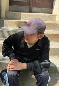 Svart skjorta med knappar och volanger vid ärmsluten, grå keps med en logotyp, och svarta byxor med ljusblå ränder som sitter på stensteg.