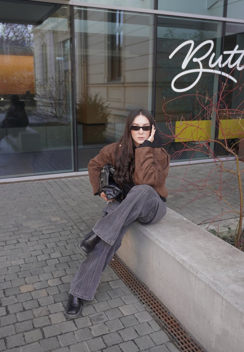 Femme en veste marron et lunettes de soleil assise sur un banc en béton devant un bâtiment en verre avec une enseigne "Butter", tenant un sac noir.