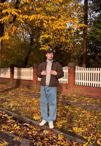 Jachetă bomber maro peste un polo gri, blugi largi albastru deschis și adidași bej. Înconjurat de frunze de toamnă și un gard alb de lemn.