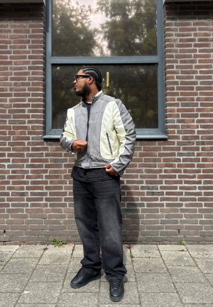 Bomber jacket gris y crema con un diseño estructurado, bolsillos con cremallera, combinada con jeans oscuros y zapatillas negras, de pie contra una pared de ladrillo.