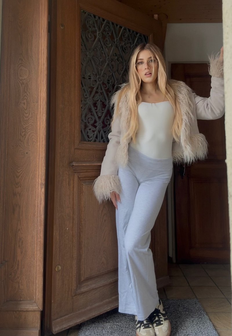 Calças cinza claro à boca de sino com um bodysuit branco e um casaco creme com detalhes em pelagem. Fundo com uma porta de madeira e um tapete texturizado.