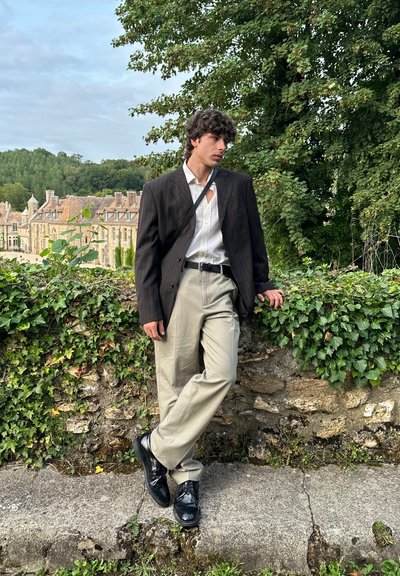 Blazer negro de rayas sobre una camisa blanca, pantalones crema y zapatos negros brillantes. De pie contra una pared de piedra con vegetación.