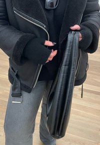 Άτομο που φοράει μαύρο δερμάτινο μπουφάν με γούνα και δαχτυλο Gloves κρατάει μαύρη δερμάτινη τσάντα, στέκεται σε ξύλινο πάτωμα.