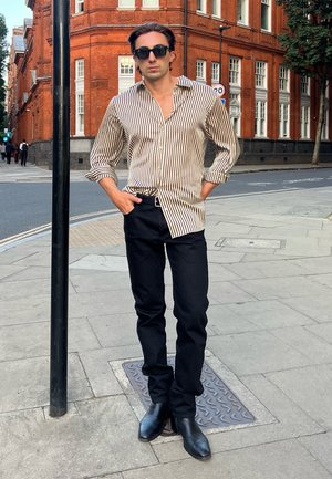 Mann mit schwarzer Sonnenbrille, gestreiftem beige-schwarzem Hemd, schwarzen Hosen und Stiefeln steht auf dem Bürgersteig einer Stadt in der Nähe eines roten Backsteingebäudes.