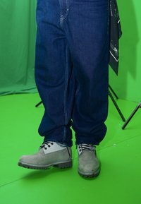 Hallid tööjalatsid mustade nööpidega, kantud koos siniste teksapükstega. Pükstel on nähtavad õmblused ja lai lõige. Taust on roheline.