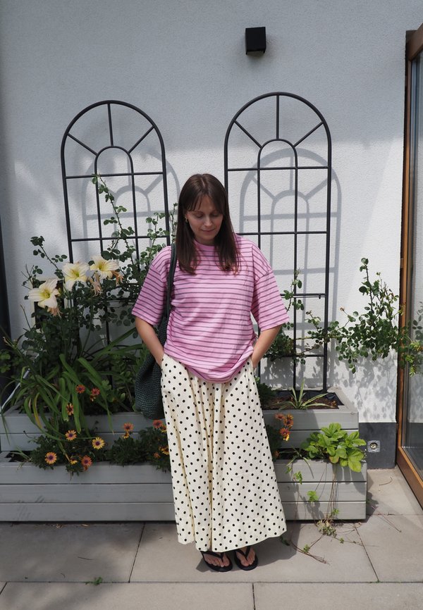 Donna con camicia rosa a righe e gonna a pois, in piedi con le mani nelle tasche davanti a fioriere e graticci in metallo su una parete bianca.