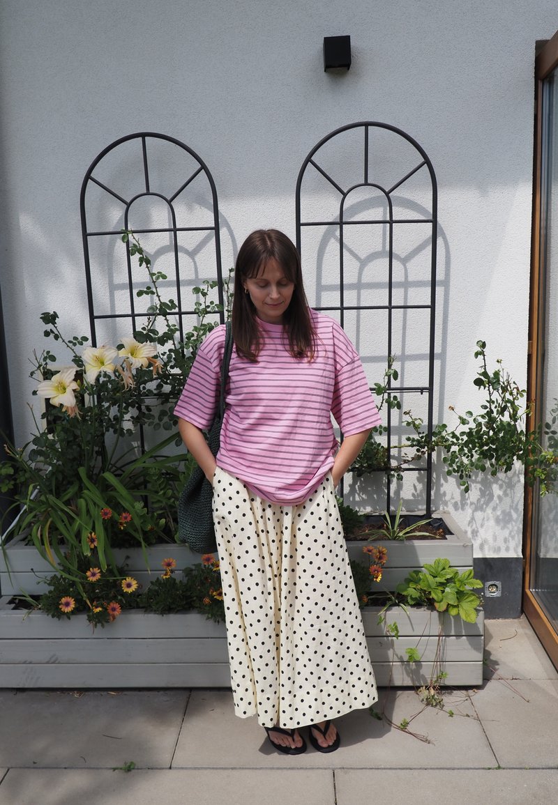 Femme en chemise rayée rose et jupe à pois, debout les mains dans les poches devant des jardinières de fleurs et des treillis métalliques sur un mur blanc.