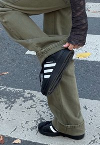 Crne Adidas Superstar tenisice s bijelim prugama, nošene s maslinasto zelenim hlačama, prikazuju teksturirani dizajn i kontrastni potplat.