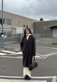 Mørkebrun, dobbelspent kåpe med store lommer, båret over en hvit turtleneck og lyse bukser. Holder en svart håndveske med sløyfedetalj.