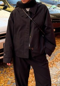 Černá, volnější bunda s knoflíkovým zapínáním a velkým límcem, doplněná odpovídajícím šátkem. Nošená přes tmavé kalhoty, na podzimním listí.