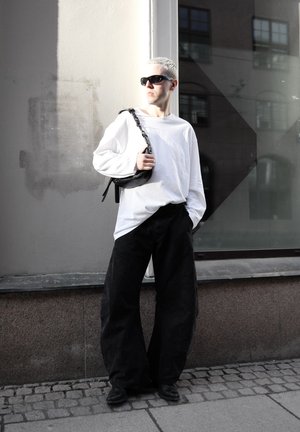 Persona con cabello corto rubio y gafas de sol lleva una camisa blanca de manga larga, pantalones negros holgados y sostiene un bolso negro al hombro junto a una pared de edificio.