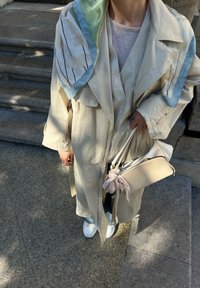 Trench coat beige con una finitura morbida e testurizzata; sciarpa con motivo verde chiaro e avorio; borsa bianca con un accento in tessuto; sneakers bianche.