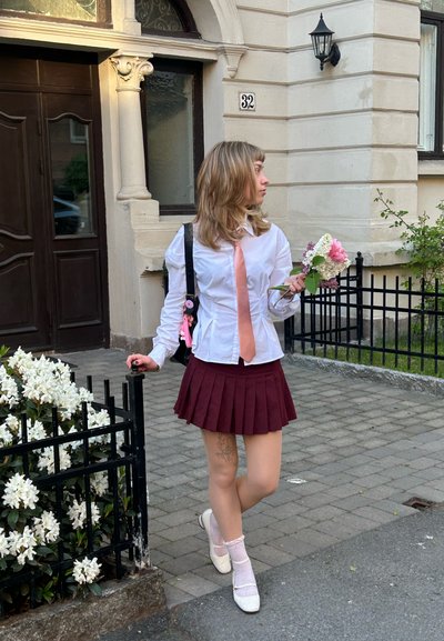 Mujer con camisa blanca, corbata rosa y falda granate sosteniendo flores, apoyada en una valla negra cerca de la entrada del edificio con el número 32.