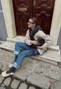 Γυναίκα με γυαλιά, μπεζ σακάκι, γκρι κασκόλ και μπλε τζιν, καθισμένη σε πέτρινες σκάλες, κρατώντας μια μαύρη τσάντα κοντά σε μια ξύλινη πόρτα.