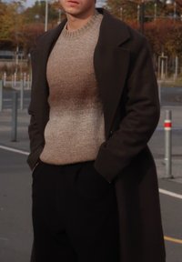 Mørk frakke over en lysebrun, struktureret sweater; begge er tætsiddende. Frakken har brede revers, og outfittet inkluderer afslappede, sorte bukser.