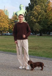 Maglione di lana marrone, pantaloni in corduroy crema, sneakers bianche. La persona tiene un guinzaglio per un bassotto marrone. Statua di cavallo verde sullo sfondo.