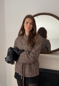 Femme aux longs cheveux bruns portant un manteau marron à motifs et des gants noirs, tenant un sac à main noir devant un miroir rond.