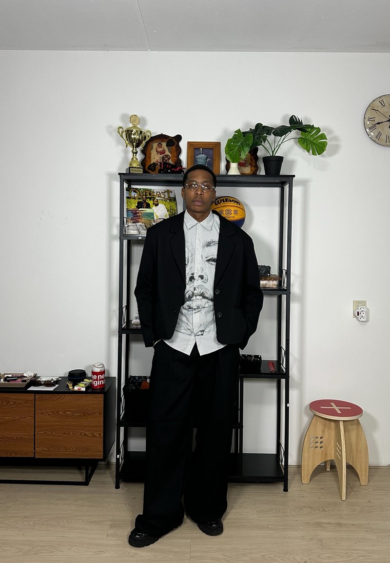 Blazer preto sobre uma camisa branca com uma estampa de rosto, combinado com calças largas pretas e sapatos pretos. Exibido em frente a uma prateleira decorada.