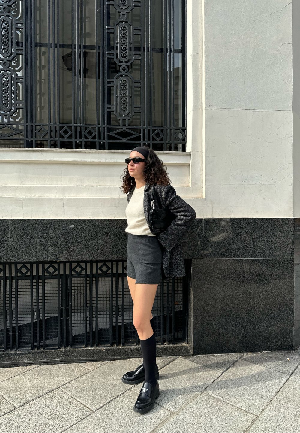 Blazer negro sobre un suéter claro, pantalones cortos grises, calcetas altas negras y zapatos negros; cabello rizado y gafas de sol, de pie contra una pared texturizada.