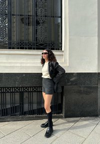 Blazer negro sobre un suéter claro, pantalones cortos grises, calcetas altas negras y zapatos negros; cabello rizado y gafas de sol, de pie contra una pared texturizada.