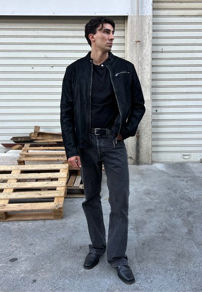 Chaqueta de cuero negra sobre una camiseta negra, combinada con jeans oscuros y zapatos negros. Los accesorios incluyen un cinturón con una hebilla de plata.