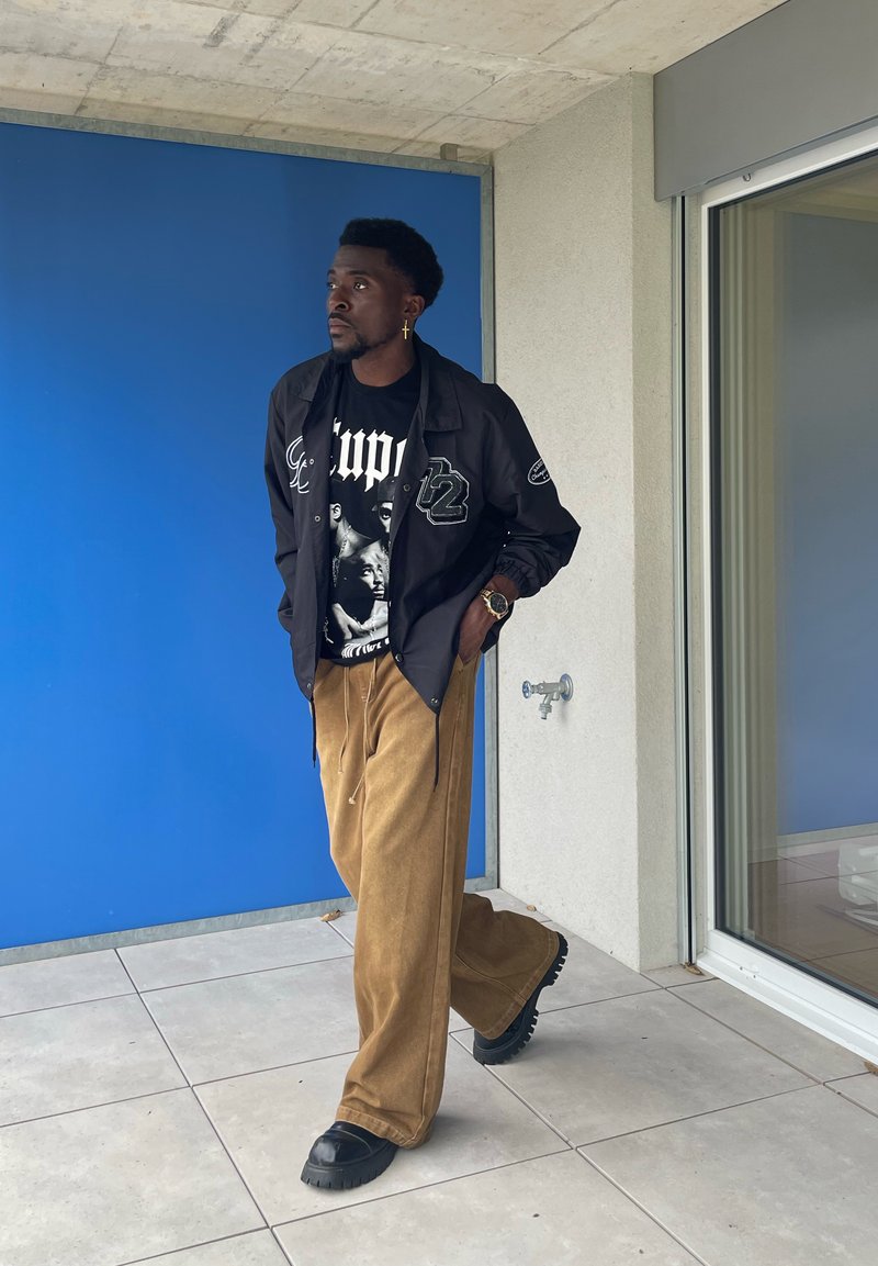 Veste noire zippée avec des accents de logo sur un T-shirt à motifs noirs, associée à un pantalon large en velours côtelé beige et des bottes noires à semelles épaisses.