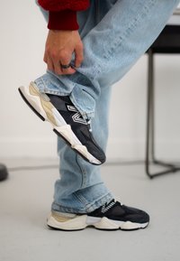 Μαύρα και κρεμ New Balance παπούτσια με μαξιλαρωτές σόλες, αξεσουάρ από ανθεκτικό δίχτυ και ανακλαστικές λεπτομέρειες, σε συνδυασμό με ανοιχτό γαλάζιο τζιν.