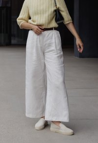 Bílé lněné kalhoty s širokými nohavicemi a texturovaným povrchem, doplněné světle žlutým pleteným svetrém a béžovými nazouvacími botami. Černá kabelka doladí celý outfit.