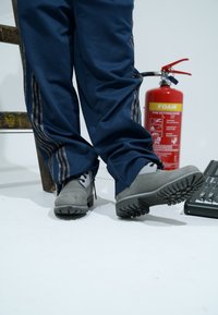 Pelēki drošības apavi ar gumijas zolēm, kopā ar tumši ziliem sporta biksēm ar teksturētām sānu svītrām. Fona attēlā redzams ugunsdzēšamais aparāts.