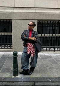 Czarna skórzana kurtka, czerwony golf, pasiasta koszula związana w pasie, luźne niebieskie jeansy, czarne buty, duża pleciona torba, ciemne okulary przeciwsłoneczne.