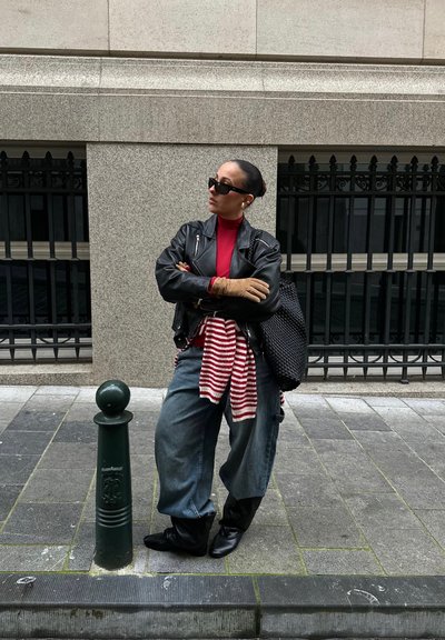 Chaqueta de cuero negra, cuello alto rojo, camisa a rayas anudada a la cintura, jeans azules holgados, botas negras, bolso grande tejido, gafas de sol oscuras.