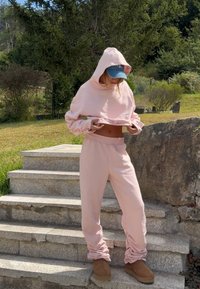 Lichtroze cropped hoodie en bijpassende joggers met elastische manchetten, gedragen met bruine enkellaarsjes, tegen een achtergrond van gras en stenen.