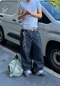 Jeans de perna larga azul-escuro com costura em contraste, combinados com uma t-shirt cinza-claro. Cinto preto com uma fivela prateada. Saco verde-oliva no chão.