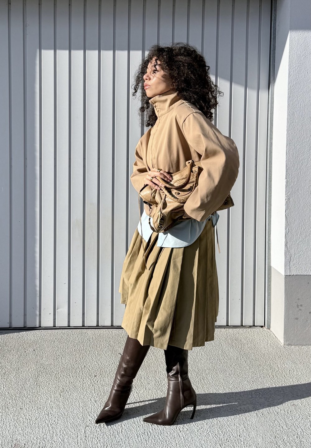 Veste beige oversize, chemise bleu clair, jupe plissée olive et bottines marron à bouts pointus. Tenant un sac marron clair avec des accents.