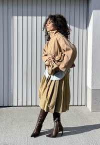 Veste beige oversize, chemise bleu clair, jupe plissée olive et bottines marron à bouts pointus. Tenant un sac marron clair avec des accents.