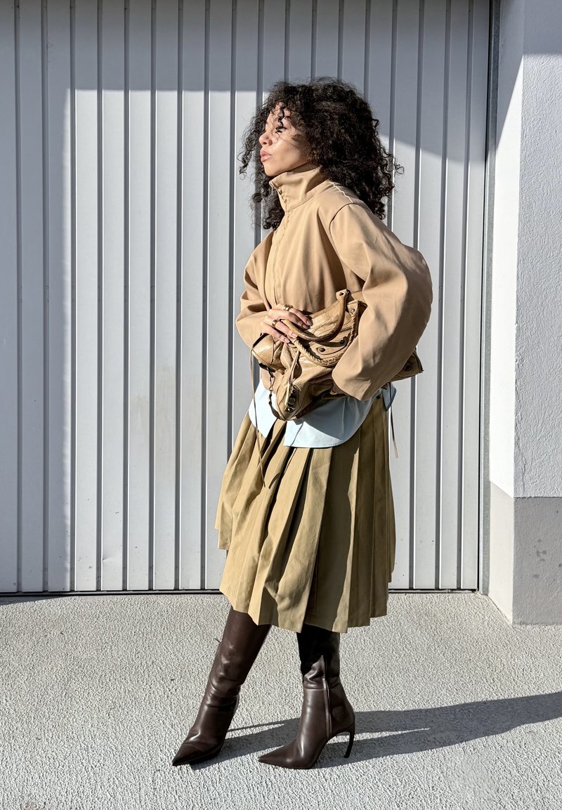Veste beige surdimensionnée, chemise bleu clair, jupe plissée olive et bottines pointues marron. Tenant un sac beige avec des accents.