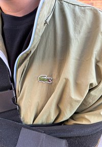 Ljusgrön, lätt jacka med dragkedja och broderad krokodil-logotyp på bröstet, lager över en svart texturerad skjorta.