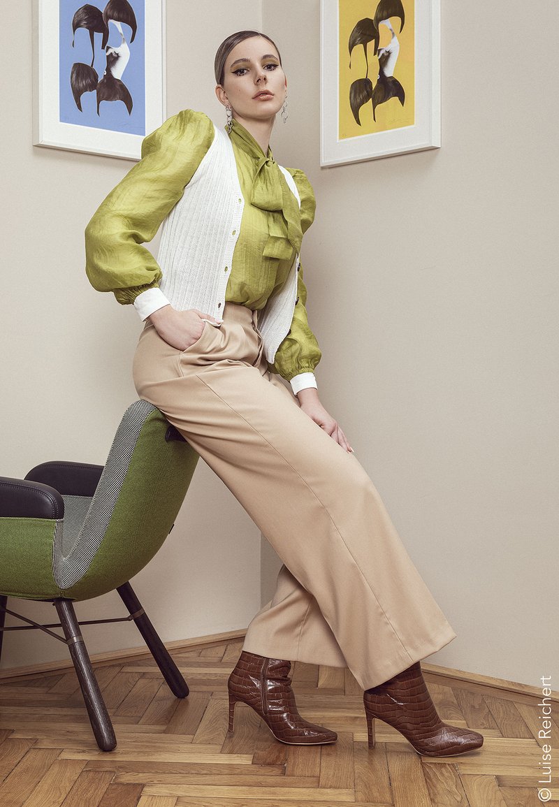 Green blouse with puffed sleeves and bow, white ribbed vest, beige trousers, and brown crocodile-embossed heeled boots; wicker chair background.