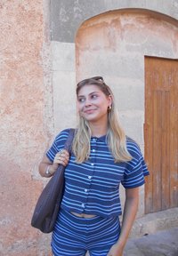 Niebieska i biała w paski krótka koszula z guziczkami i pasujące szorty. Nosi brązową plecioną torbę. W tle widać rustykalną ścianę i drzwi.