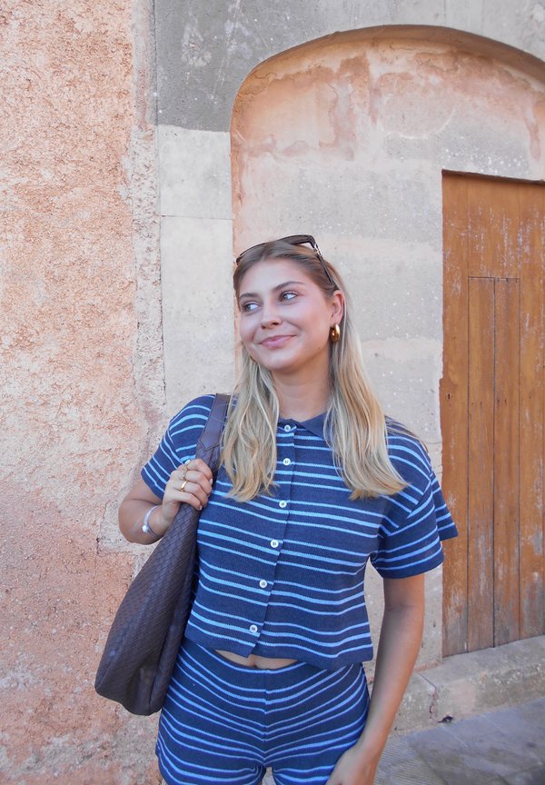 Top a maniche corte blu e bianche a righe con pantaloni corti abbinati. Porta una borsa intrecciata marrone. Lo sfondo presenta un muro e una porta in stile rustico.